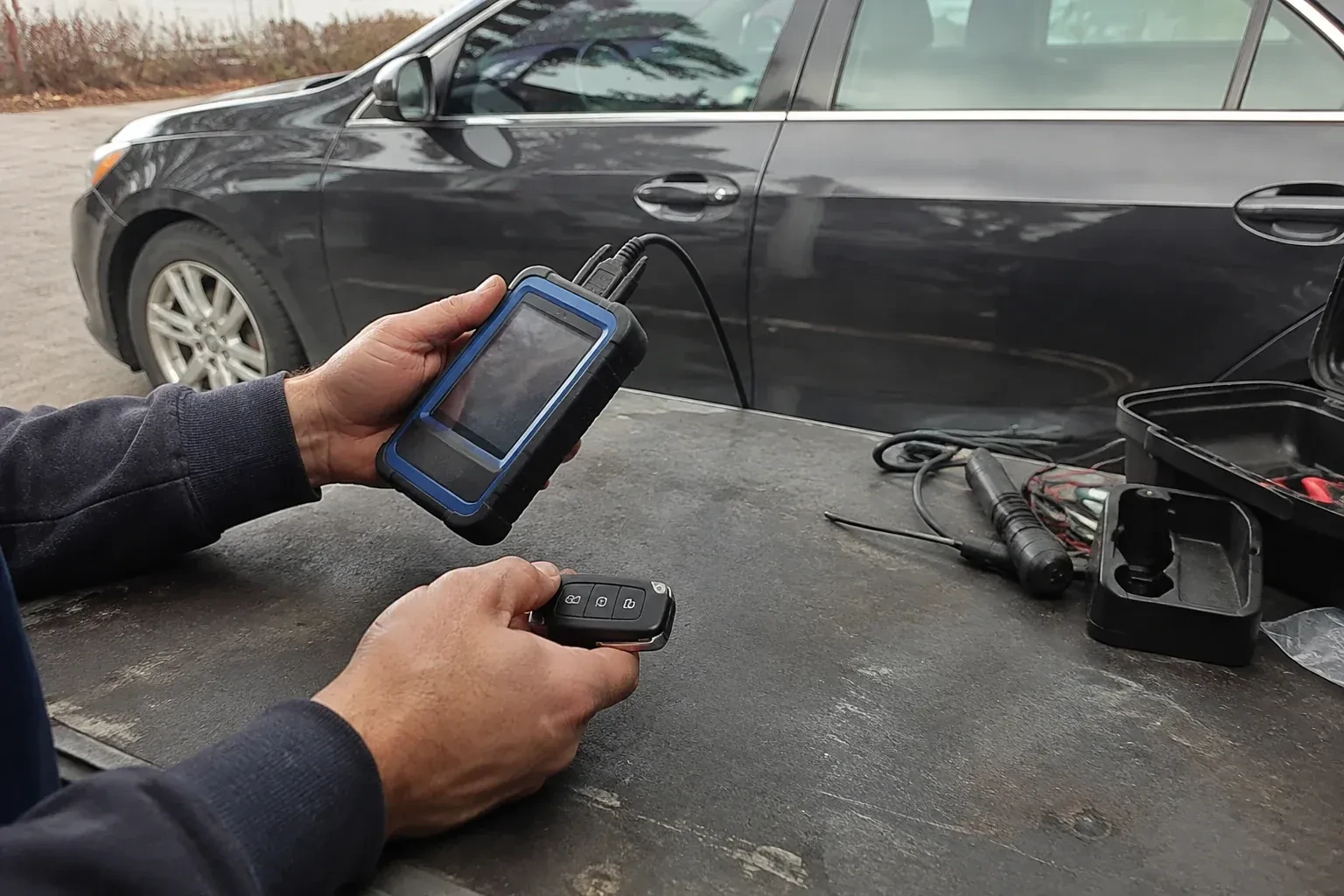 Hands holding a blue-edged electronic device and car-key fob at a worktable beside a parked car. — Transponder Key Programming, Newmarket